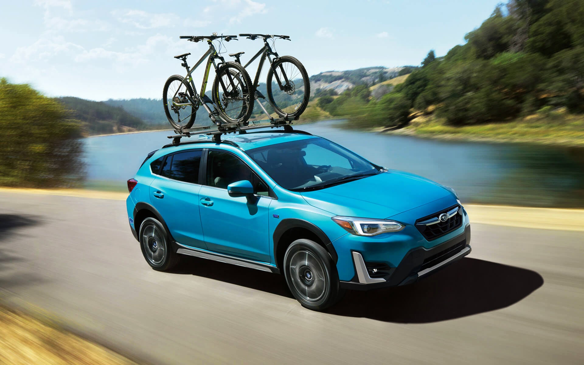 A blue Crosstrek Hybrid with two bicycles on its roof rack driving beside a river | Subaru of Spartanburg in Spartanburg SC