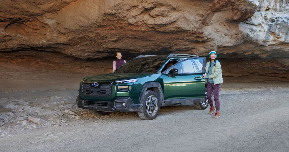 The new Subaru Outback is parked near a rocky overhang with the driver and passenger standing outside of it and smiling